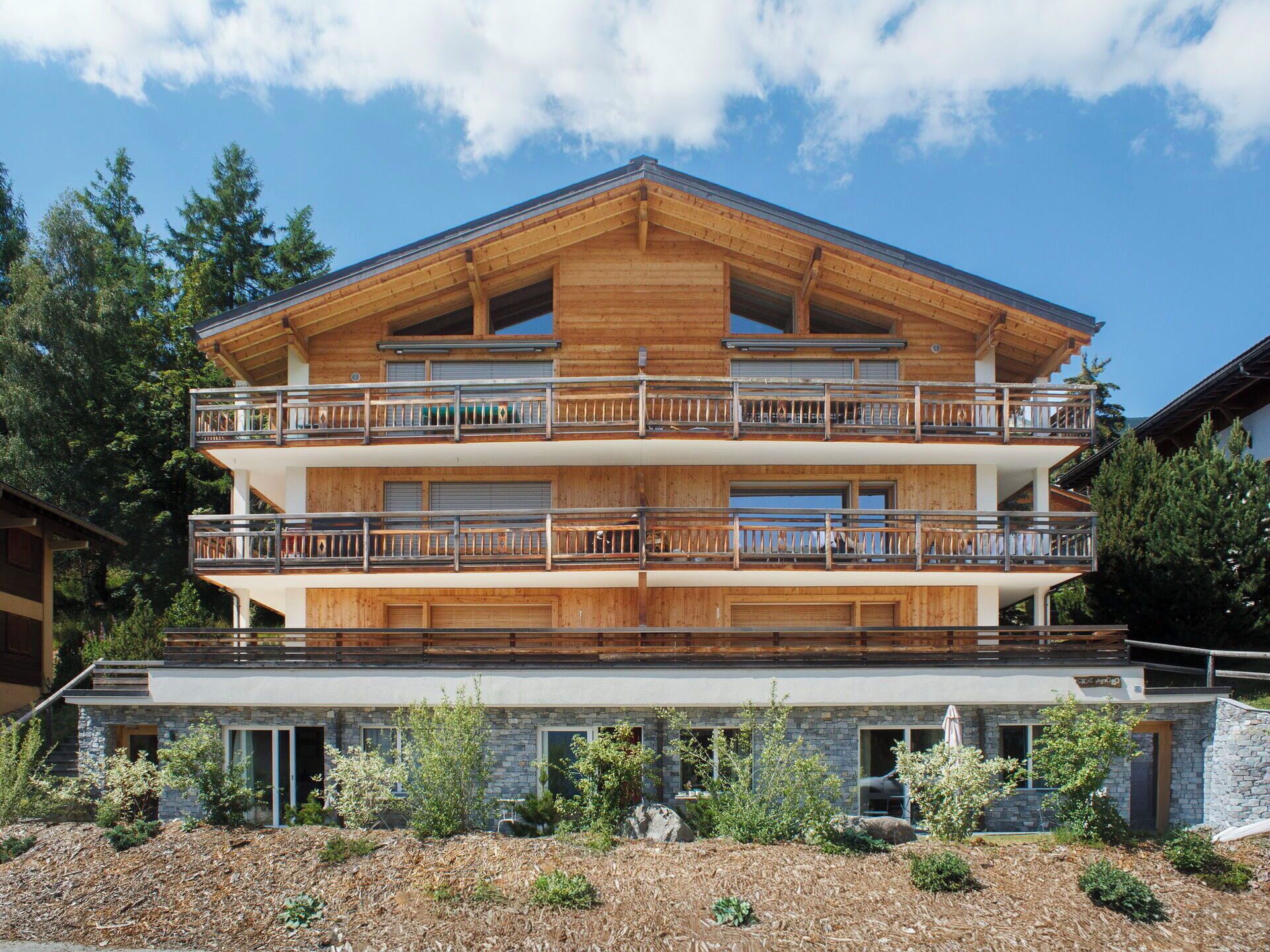 A multi-story wooden chalet with balconies, stone foundation, and surrounding vegetation.
