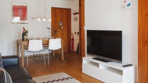 A living room with a flat-screen TV on a white stand, a black leather sofa, a dining table with chairs, and a wooden floor.