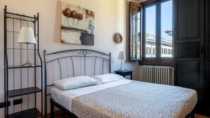 Une chambre à coucher avec un lit, une table de chevet avec une lampe, une étagère fixée au mur et une fenêtre donnant sur des immeubles.