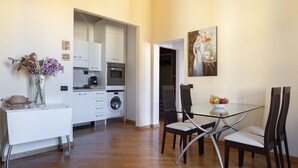 Une cuisine compacte équipée d’un four à micro-ondes, d’un four et d’un lave-linge. On y trouve une table à manger avec des chaises, ainsi qu’un vase rempli de fleurs posé sur une petite table.