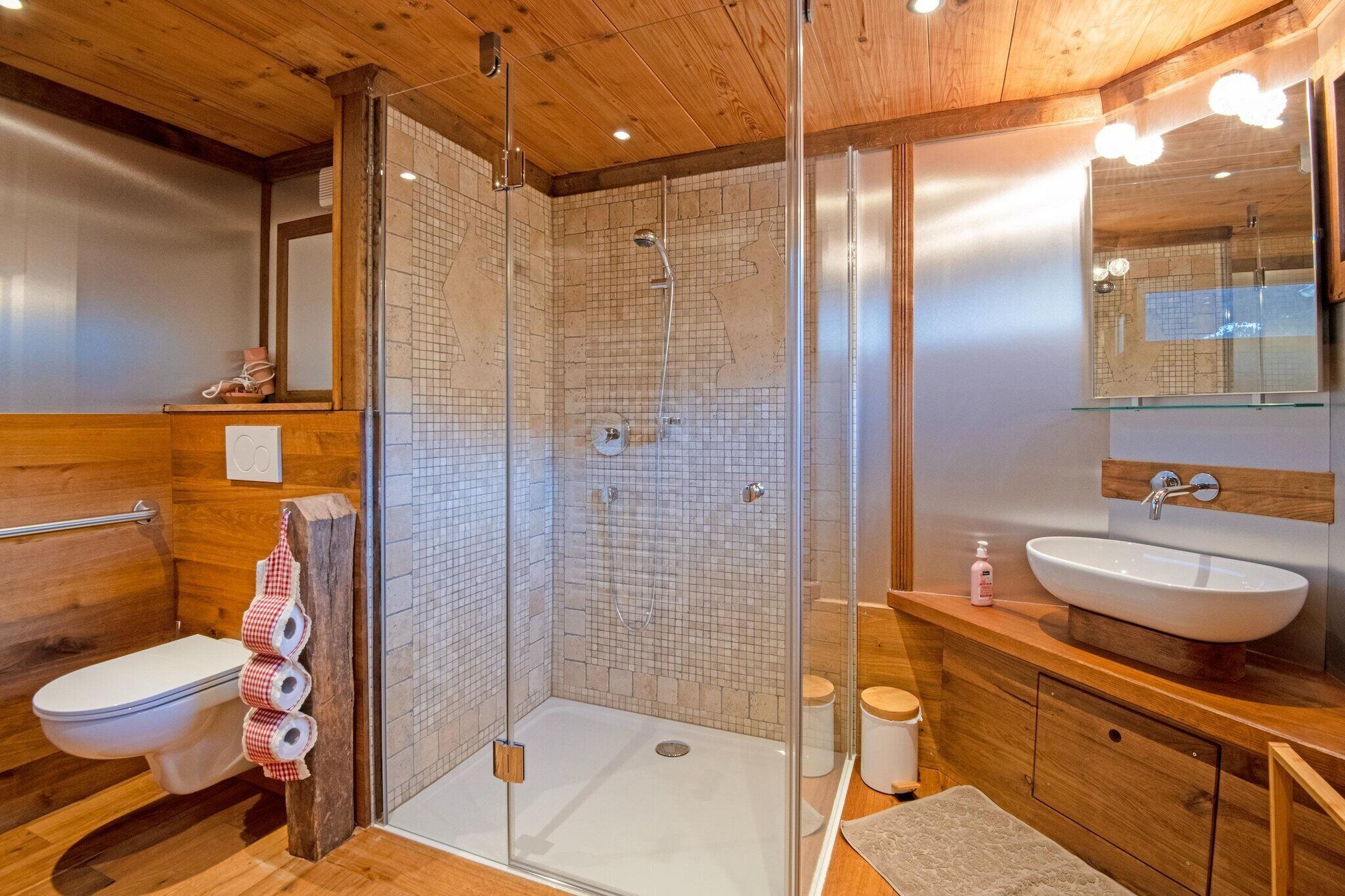 Un baño moderno con ducha acristalada, suelo de madera, lavamanos blanco y espejo sobre el lavamanos.