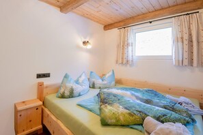 Ein hölzernes Bett mit grüner Bettdecke und blauen Kissen, ein Nachttisch, eine Wandleuchte und ein Fenster mit gemusterten Vorhängen.
