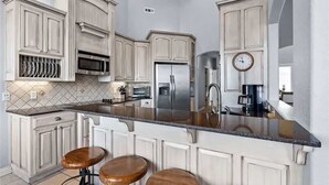 Una cocina con muebles blancos, un refrigerador de acero inoxidable, un microondas y un reloj en la pared. Hay tres taburetes de madera en la barra.