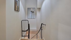 Un couloir étroit aux murs blancs et au plancher en bois. De chaque côté de l’escalier, on trouve deux rampes en métal noir.