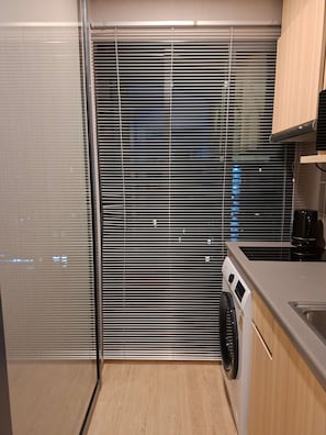 Una cocina compacta con lavadora, cafetera y vistas a través de una ventana con persianas.