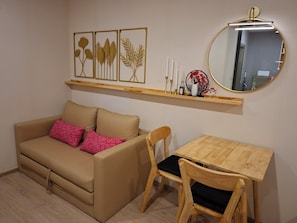 A room with a beige sofa, two wooden chairs, a small wooden table, a mirror, and wall-mounted decorations. - Ideo O2 Resort Condo BTS Bangna (Bangkok​)