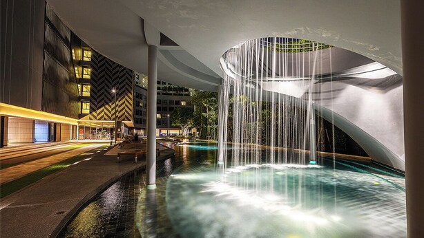 Un área de piscina interior iluminada de noche, con una cascada, rodeada de arquitectura moderna y un sendero bien iluminado.