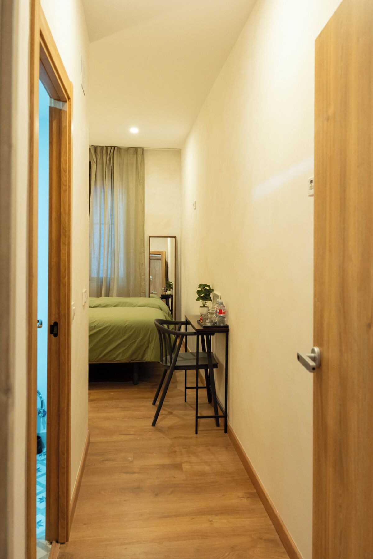 Un pasillo estrecho con piso de madera que conduce a un dormitorio con una cama y una mesa con una planta.