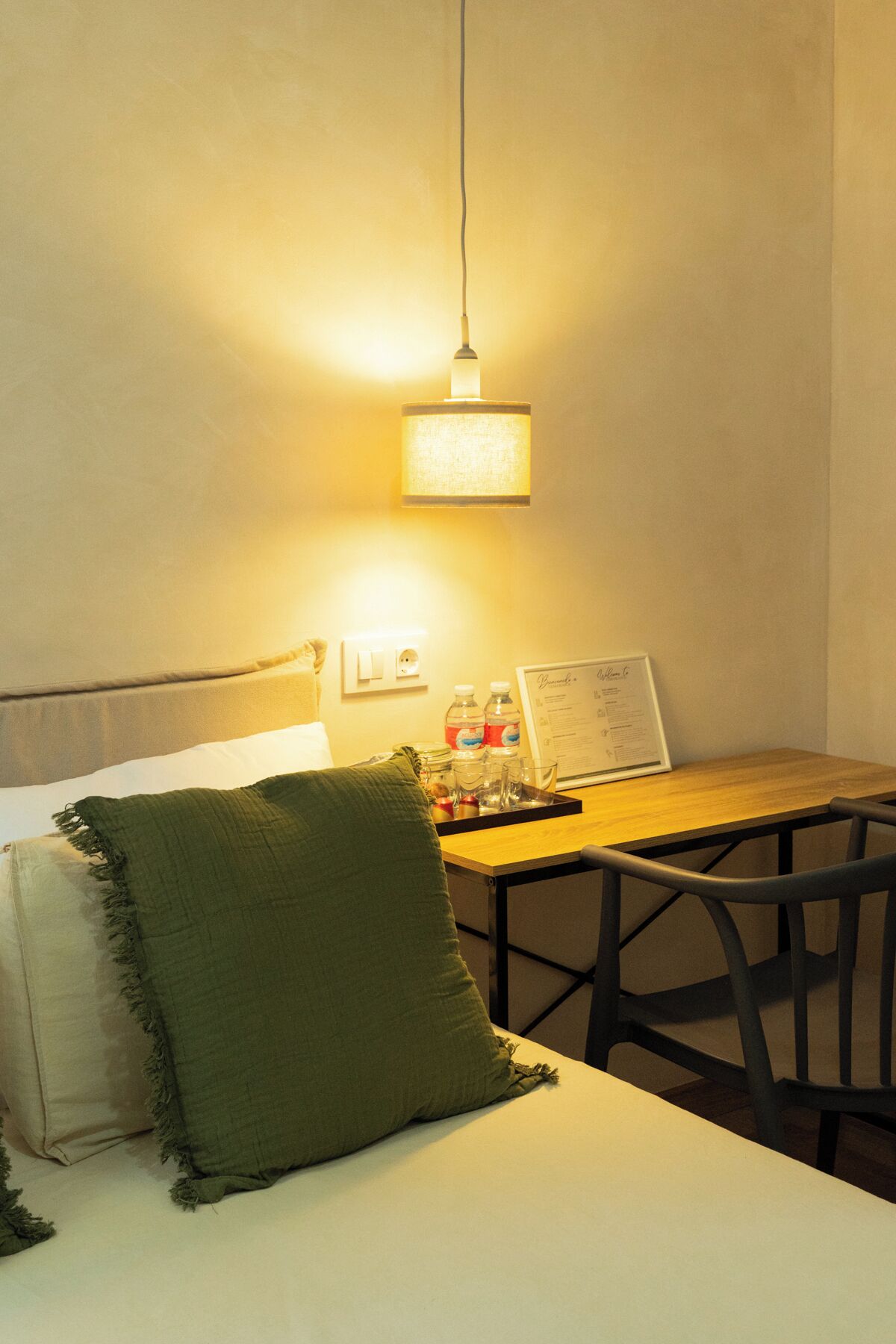 Habitación de hotel con una cama, una mesa de madera con un menú, un cojín verde y una lámpara colgante.