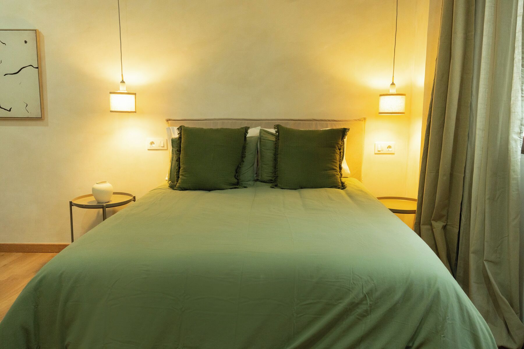 Una cama bien hecha con ropa de cama verde, dos lámparas de noche y una mesita de noche pequeña con un jarrón blanco.