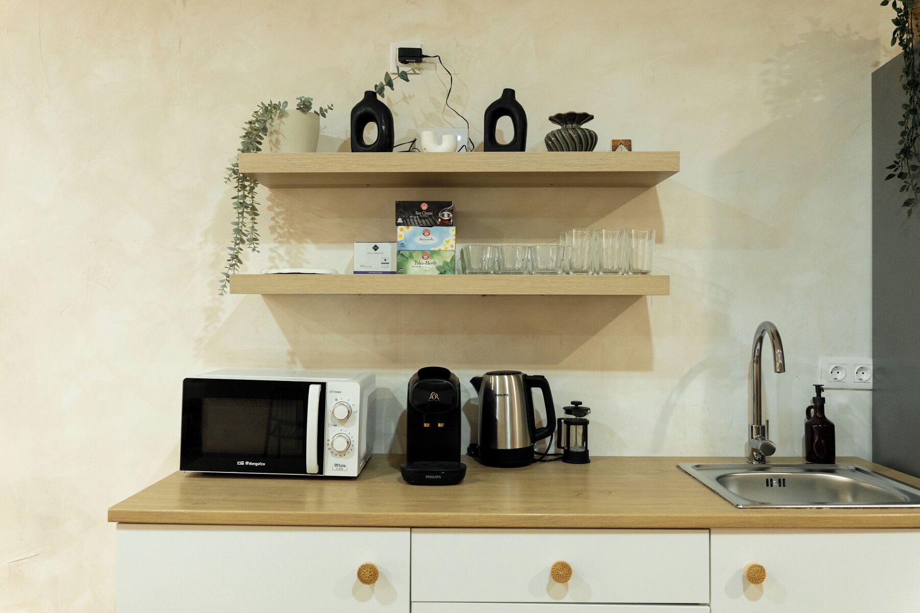 A kitchen with a microwave, coffee maker, kettle, and a sink. There are shelves above the counter holding various items.