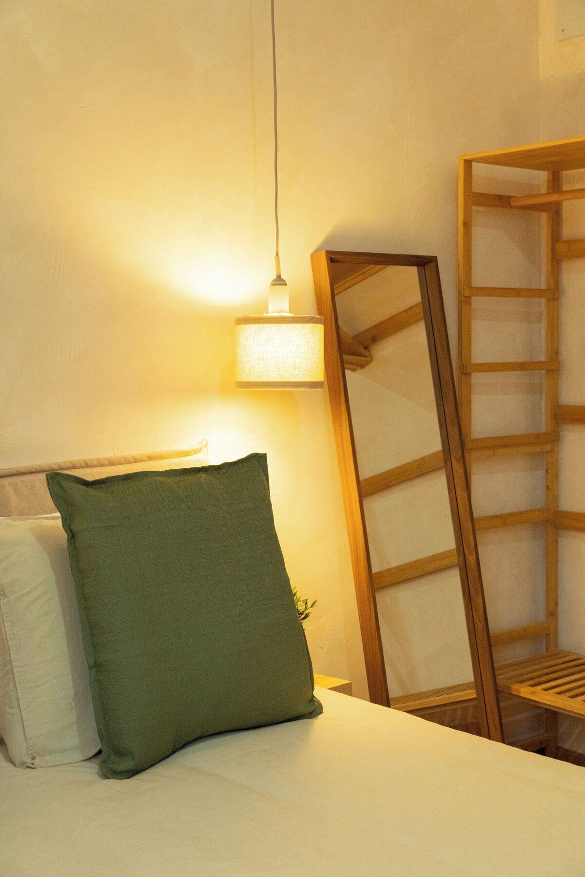 Una cama bien hecha con un almohadón verde y uno blanco, una escalera de madera apoyada en la pared, y una lámpara colgante que ilumina el espacio.