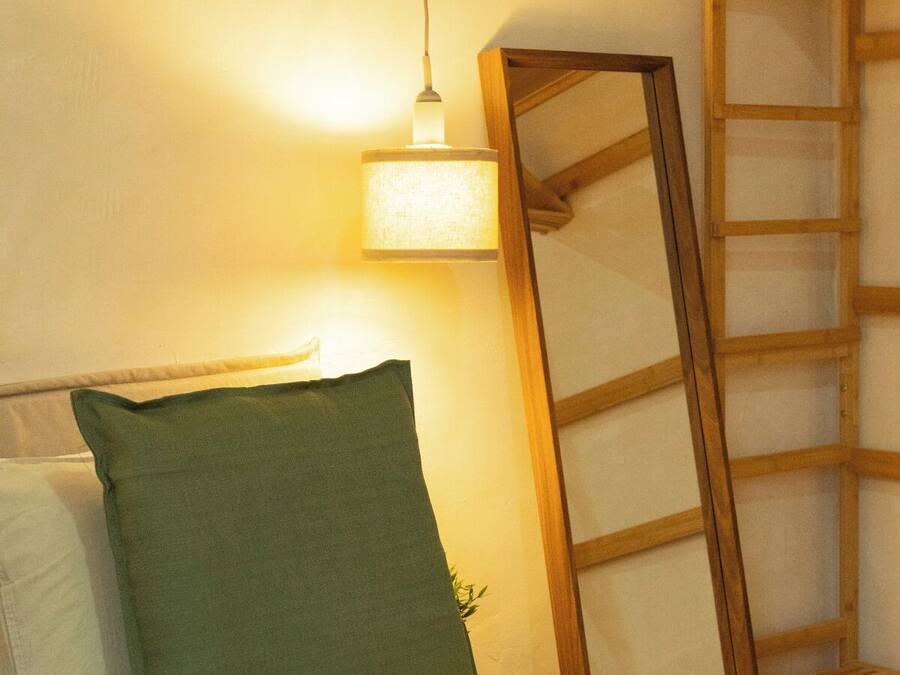 Una cama bien hecha con un almohadón verde y uno blanco, una escalera de madera apoyada en la pared, y una lámpara colgante que ilumina el espacio.