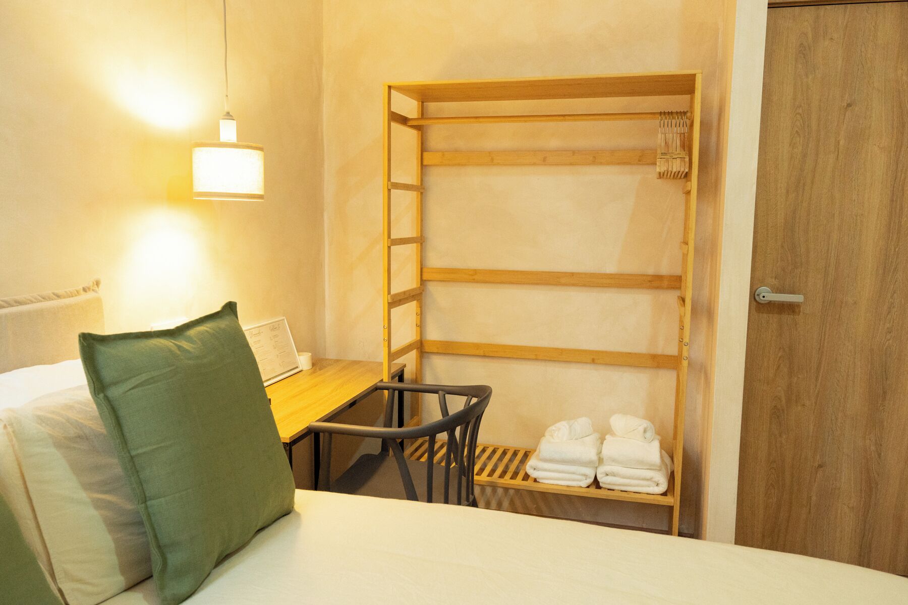Une chambre d’hôtel avec un lit superposé, un petit bureau avec une chaise, une étagère en bois avec des serviettes et une porte en bois.