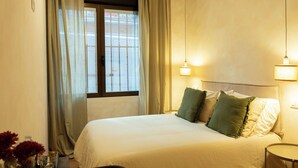 Une chambre d’hôtel avec un lit, deux lampes de chevet, une fenêtre avec des rideaux et une petite table sur laquelle est posé un vase de fleurs.