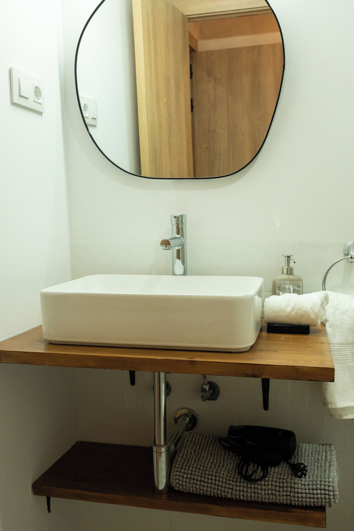 A bathroom with a white sink, a round mirror, and a wooden shelf below.