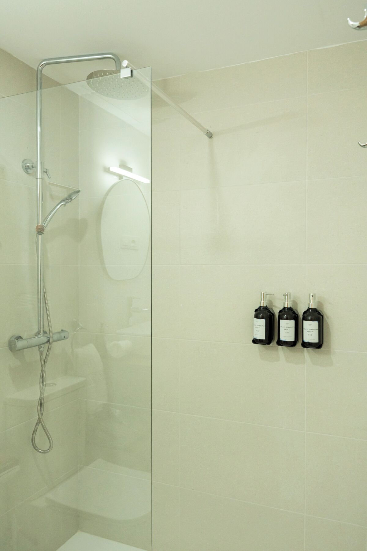 Ducha envuelta en vidrio, con cabezal de ducha de mano y llave montada en la pared. Se observan tres botellas de champú en la pared.