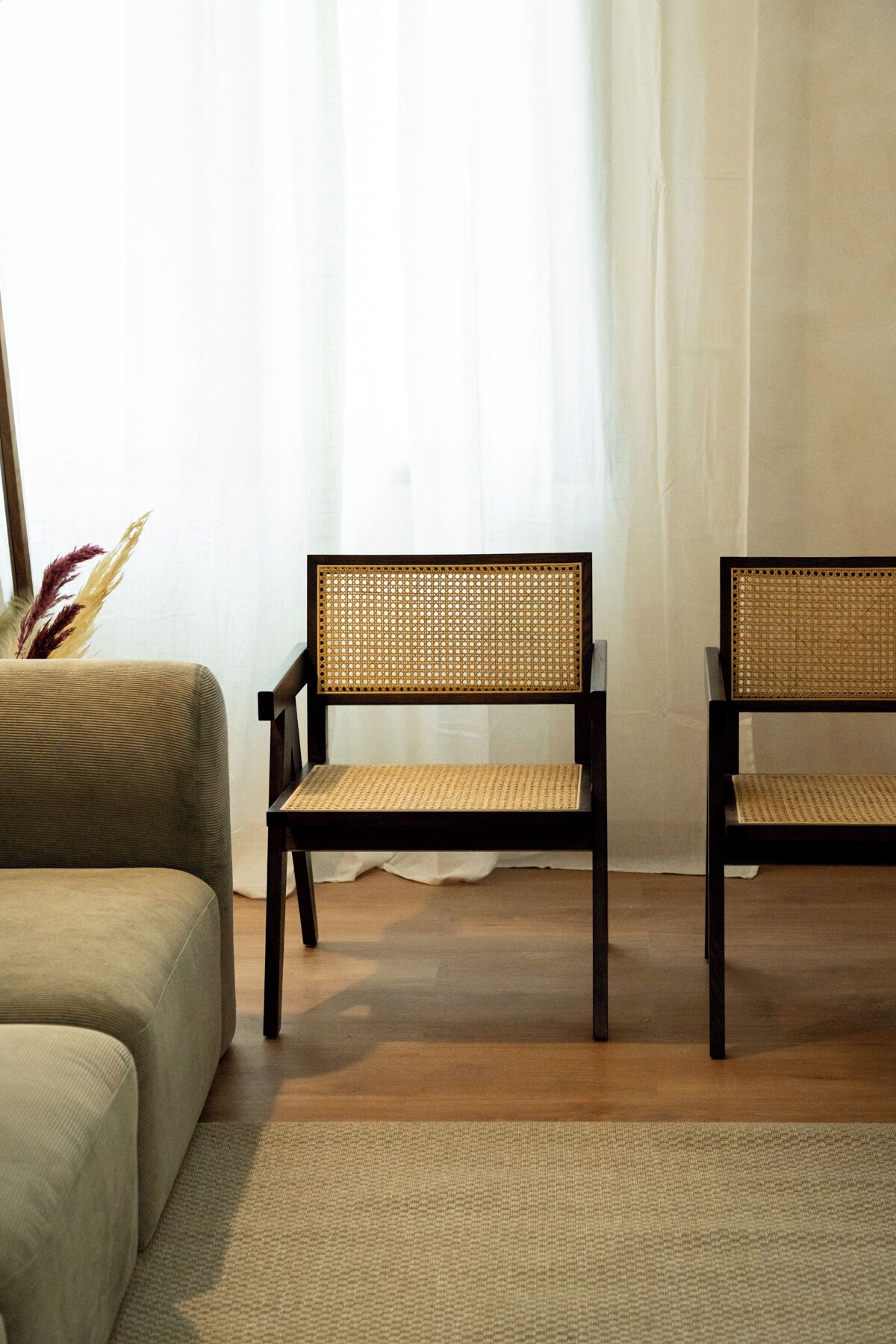 Ein Zimmer mit zwei dunklen Holzstühlen, die gewebte Sitzflächen und Rückenlehnen haben, einem beigefarbenen Sofa und einem hellen Teppich.