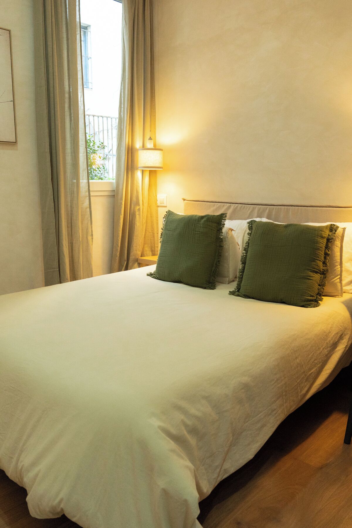 Una cama bien hecha con una colcha blanca y dos almohadas verdes, una lámpara de noche y una ventana con cortinas.