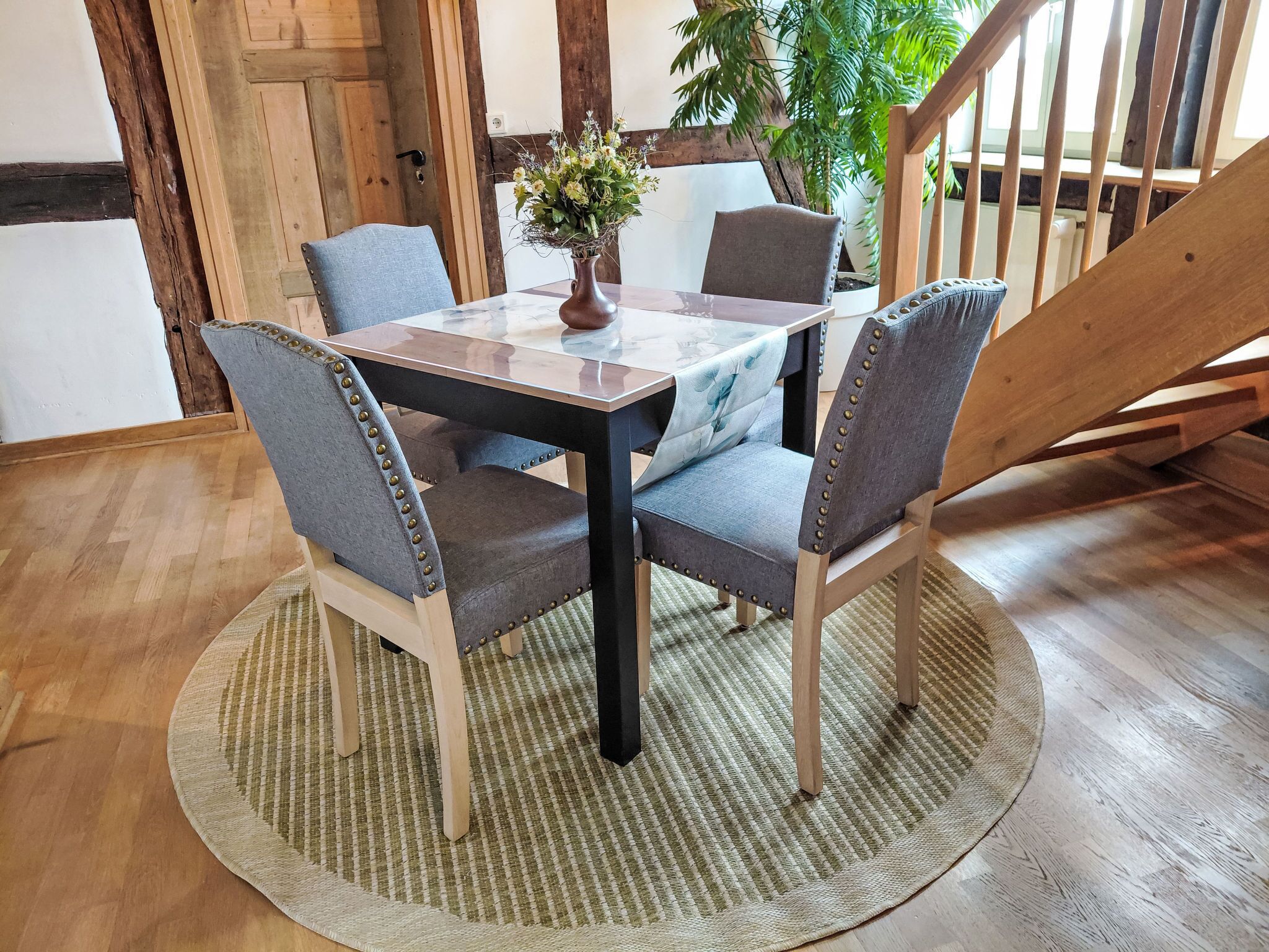 A dining area with a wooden table and chairs, a woven rug, and a potted plant.