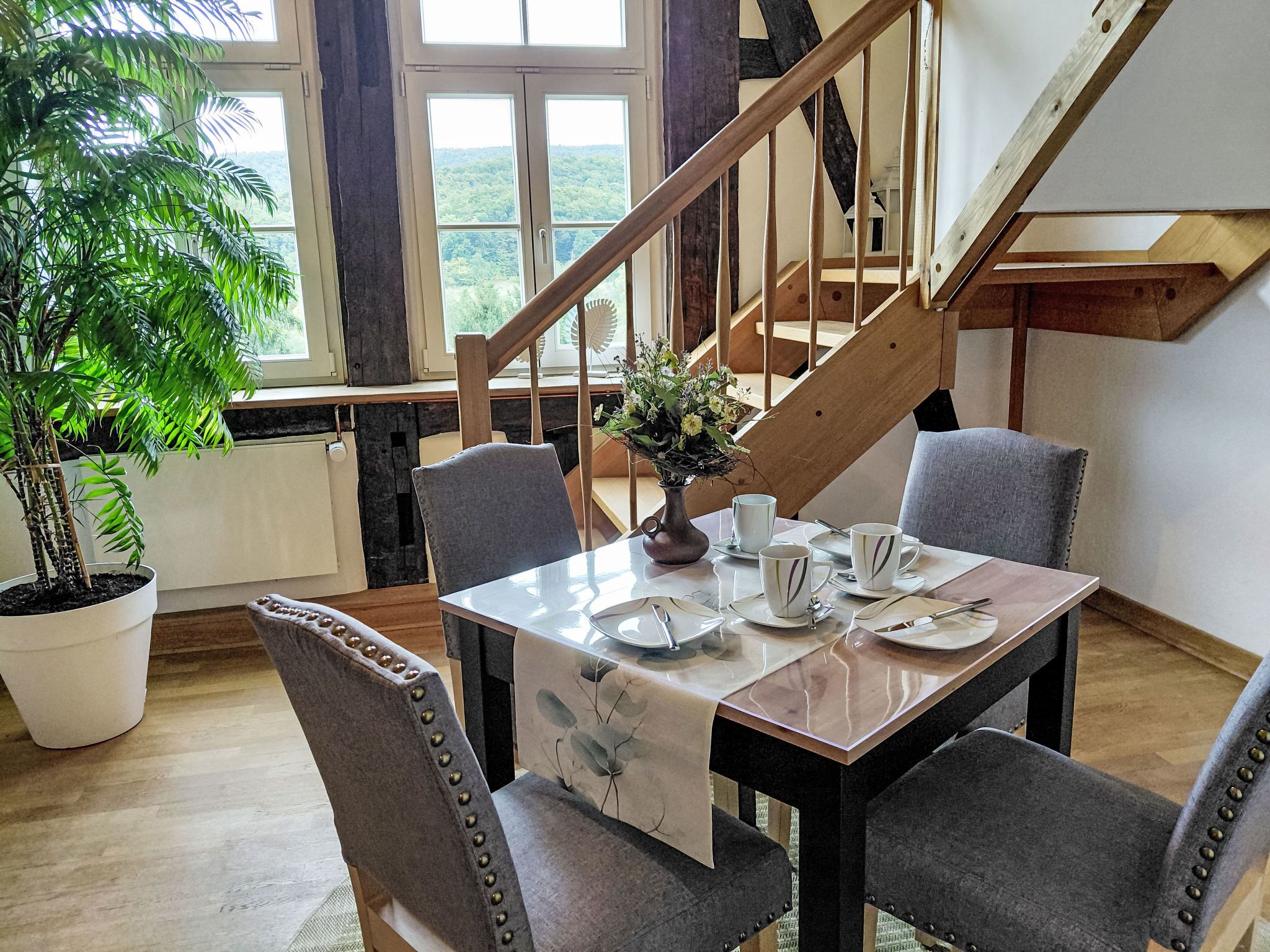 A dining area with a table set for four, surrounded by chairs with decorative studs. A potted plant is visible to the left, and a staircase leads to an upper level.