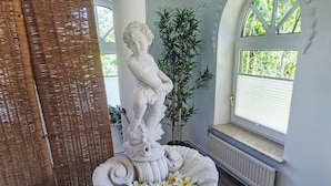 A statue of a child on a pedestal with flowers, a wicker chair, and a window with a view of greenery.