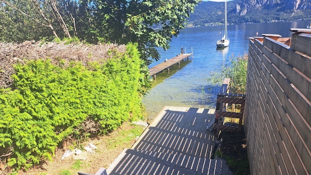 A wooden deck with a bench overlooking a calm river, surrounded by greenery and a wooden fence.
