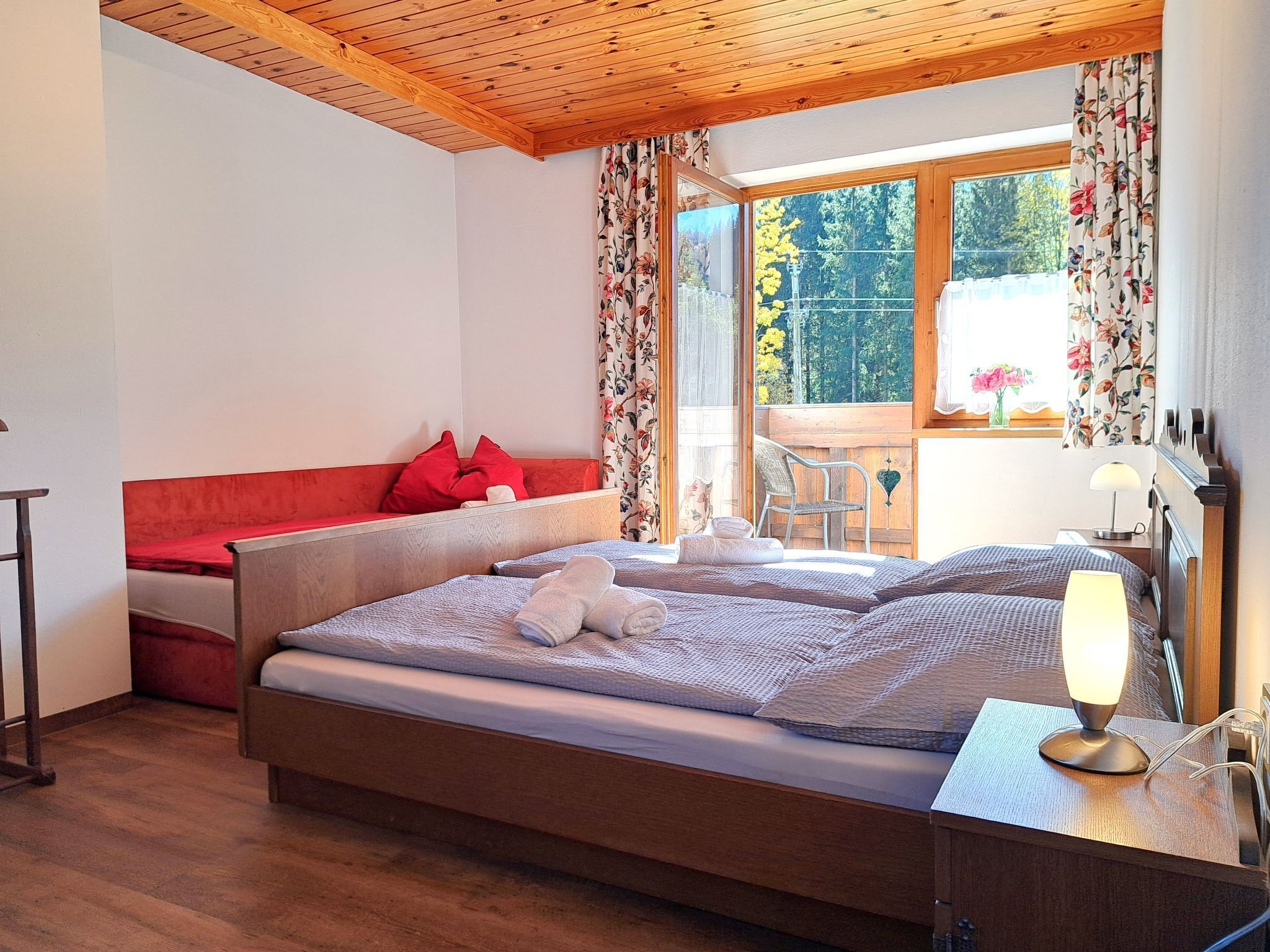 A bedroom with a wooden ceiling, a bed with a red headboard, a red sofa, a chair, a table with a lamp, and a window with floral curtains.