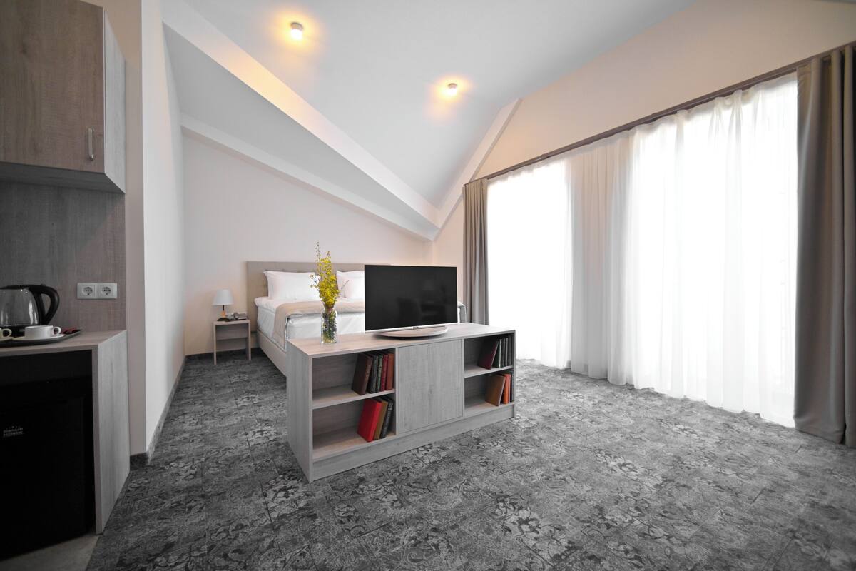 A modern hotel room with a loft bed, a flat-screen TV, a built-in bookshelf, and a large window with curtains.