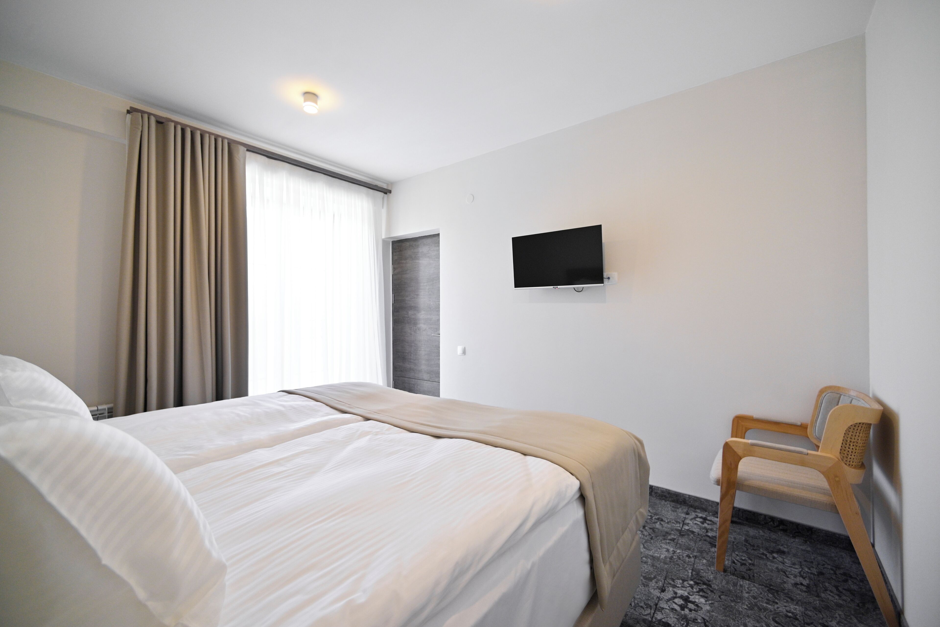 Quarto de hotel com cama, televisão, poltrona e janela com cortinas.