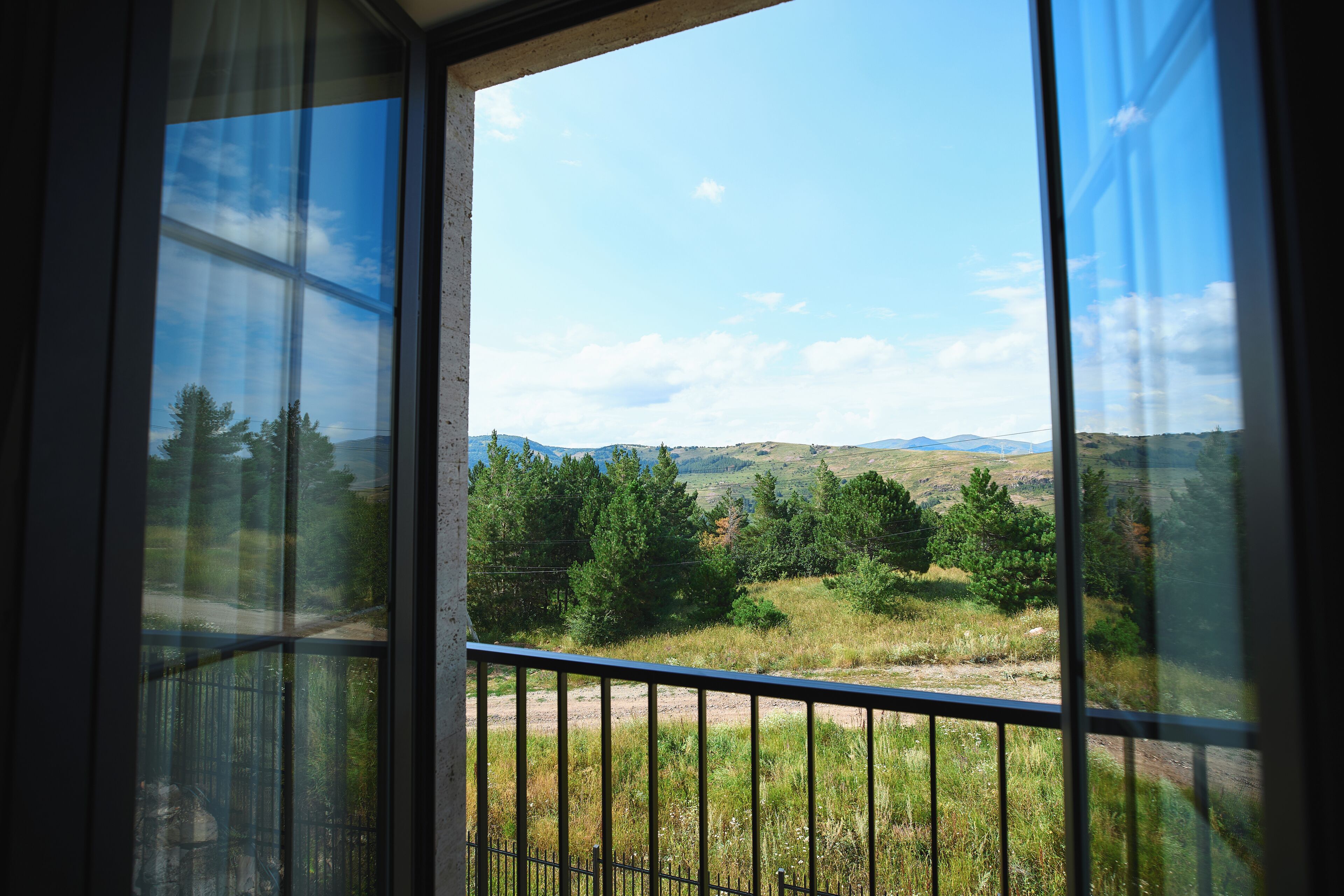 A view from a window with a metal railing, overlooking a landscape with trees and hills.