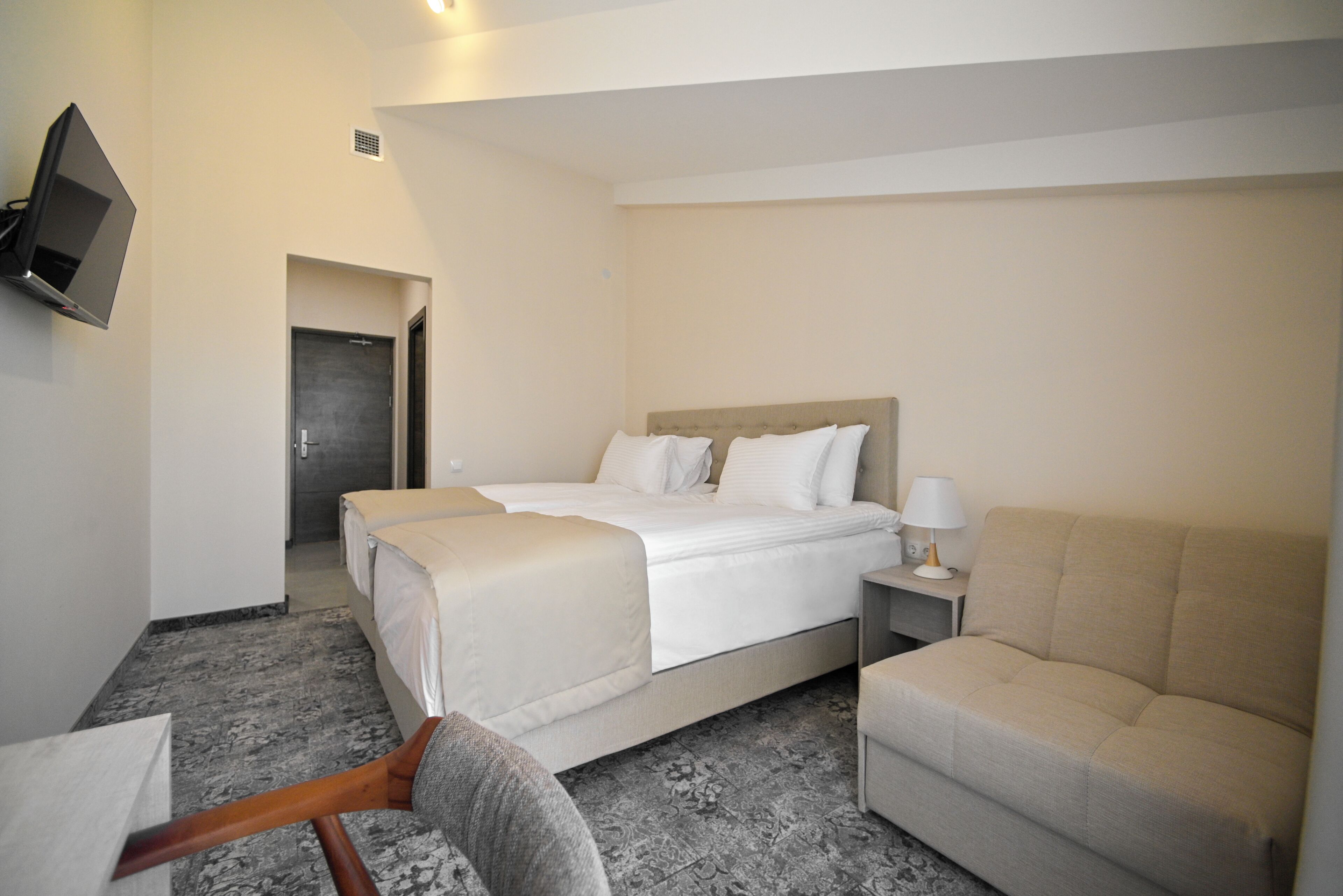 A hotel room with a bed, a sofa, a TV mounted on the wall, and a bedside table with a lamp.
