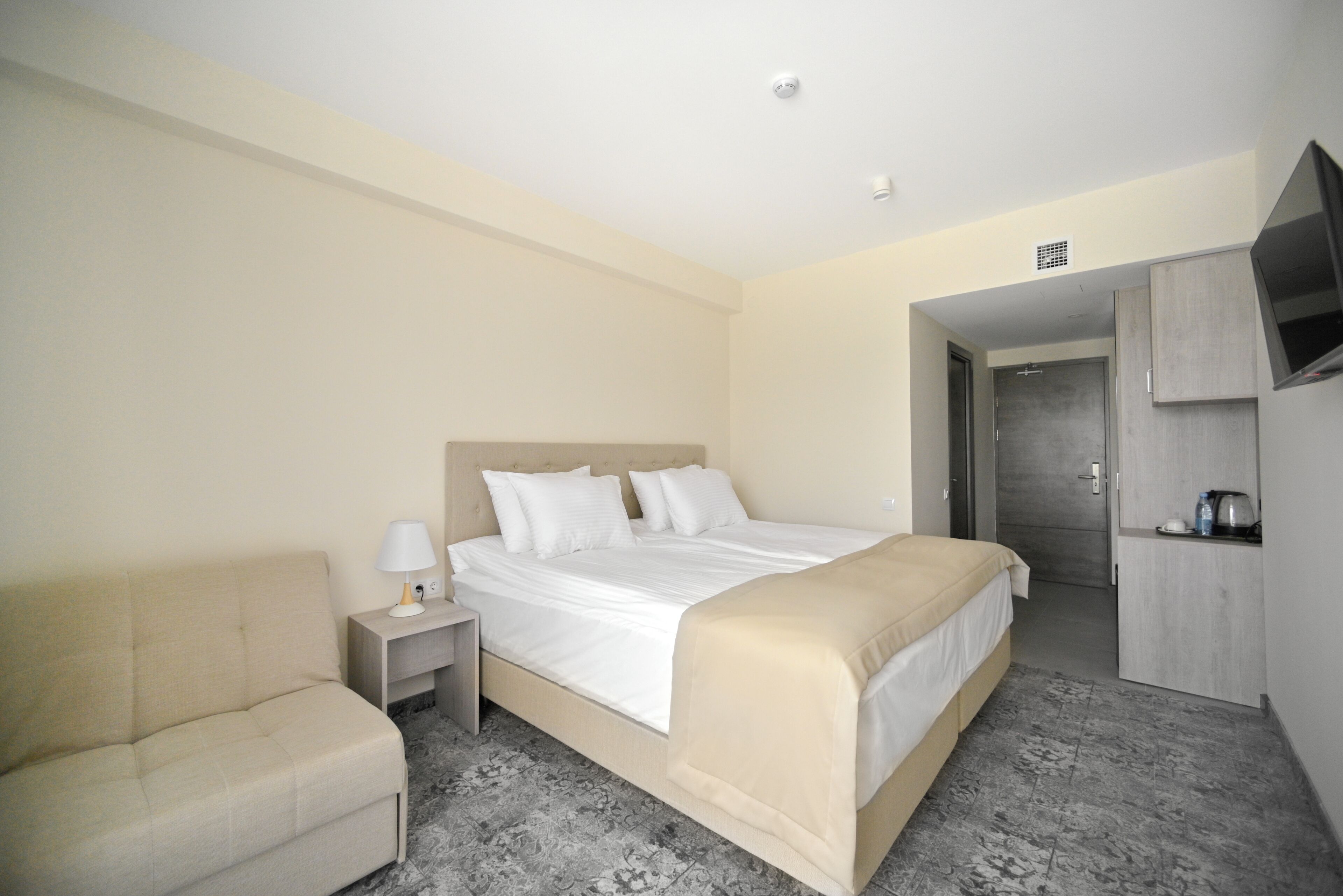 Una habitación de hotel con una cama, una mesita de noche con lámpara, un sofá y un televisor en la pared.