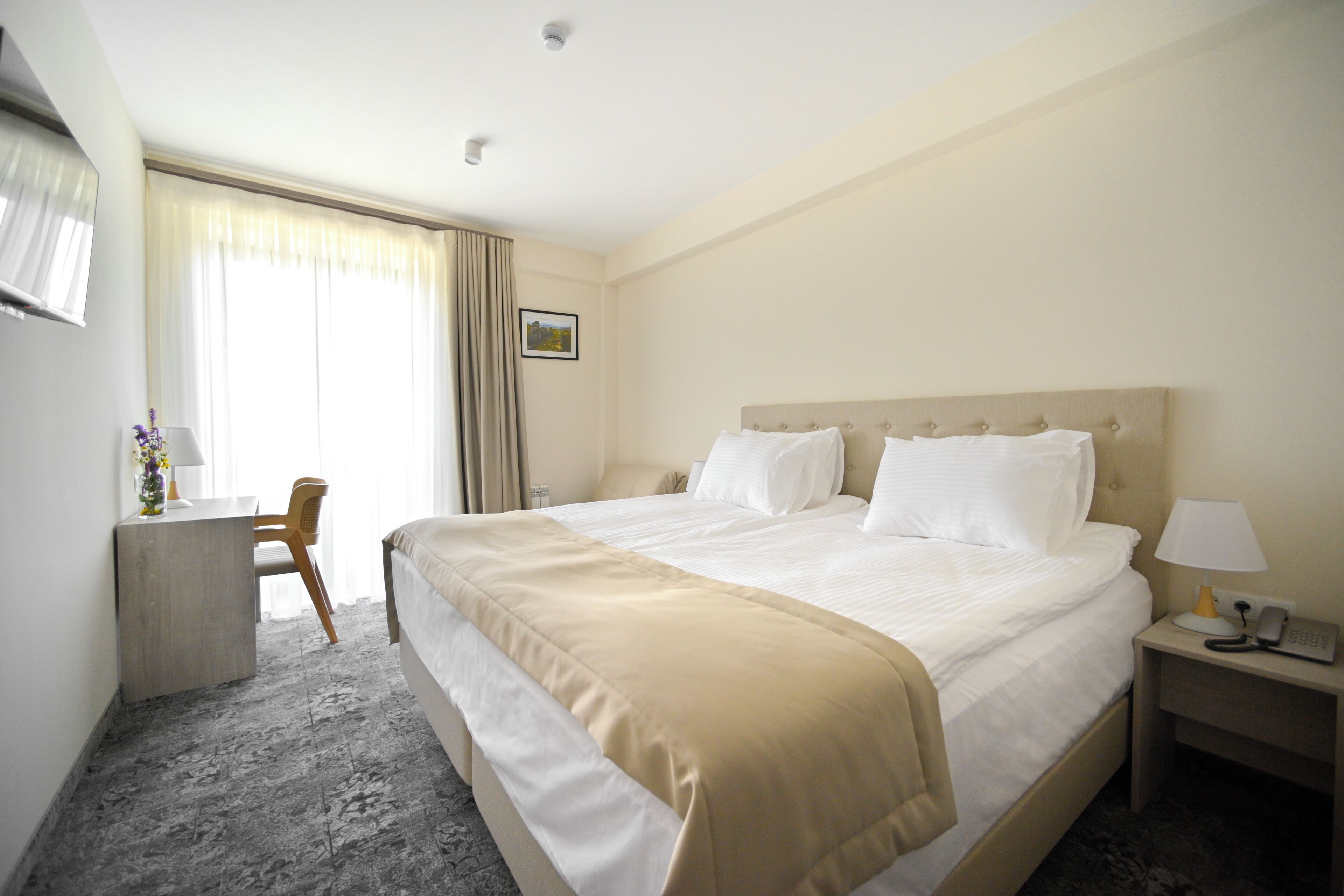 A hotel room with a large bed, a desk, a chair, and a nightstand with a lamp.