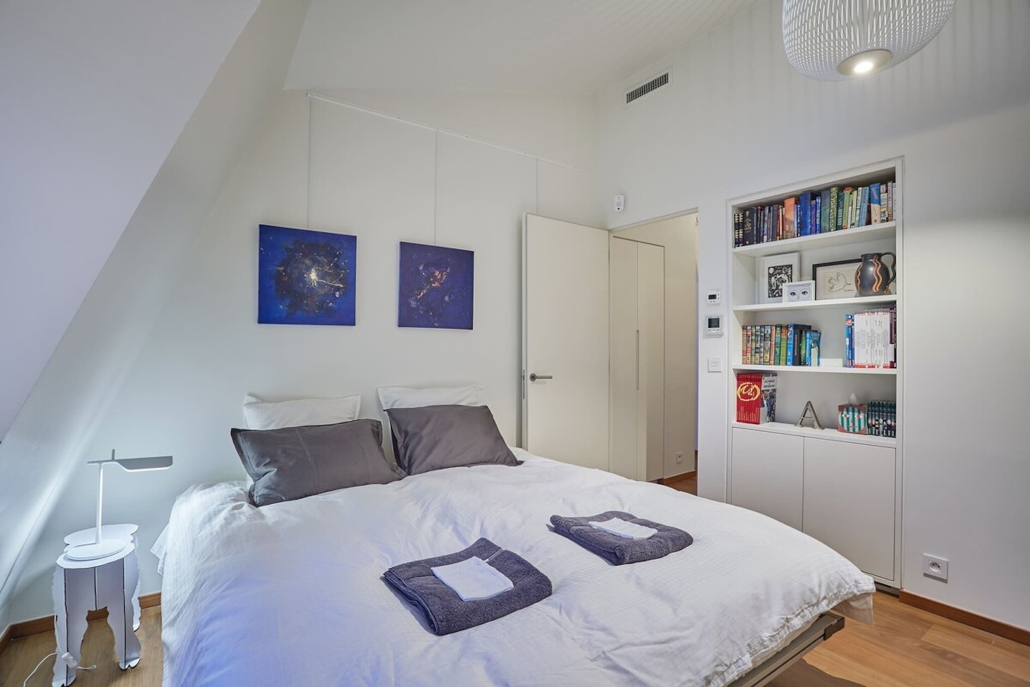Ein Schlafzimmer mit einem Bett, zwei Kissen, einem Nachttisch, einer Leselampe, einem Bücherregal und zwei Wandbildern.