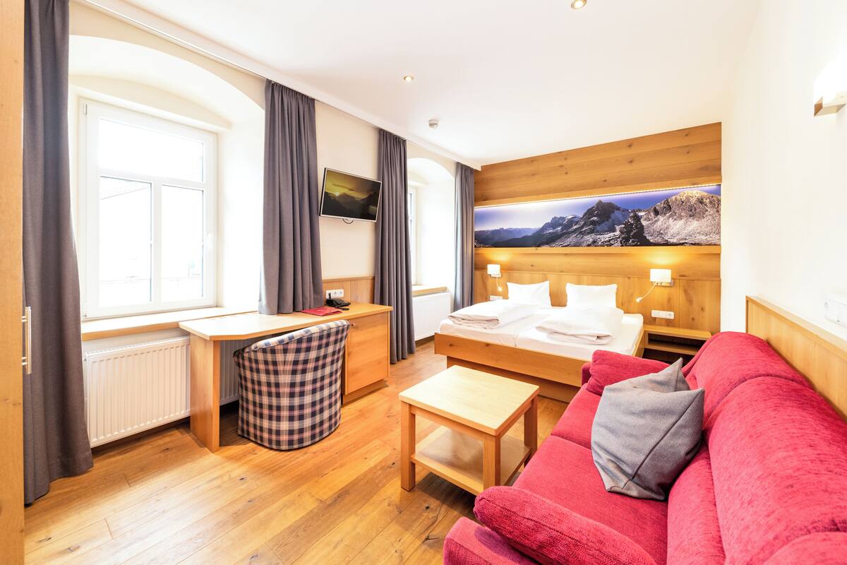 A hotel room with a wooden bed, a red sofa, a desk with a chair, a small table, and a large window with curtains.