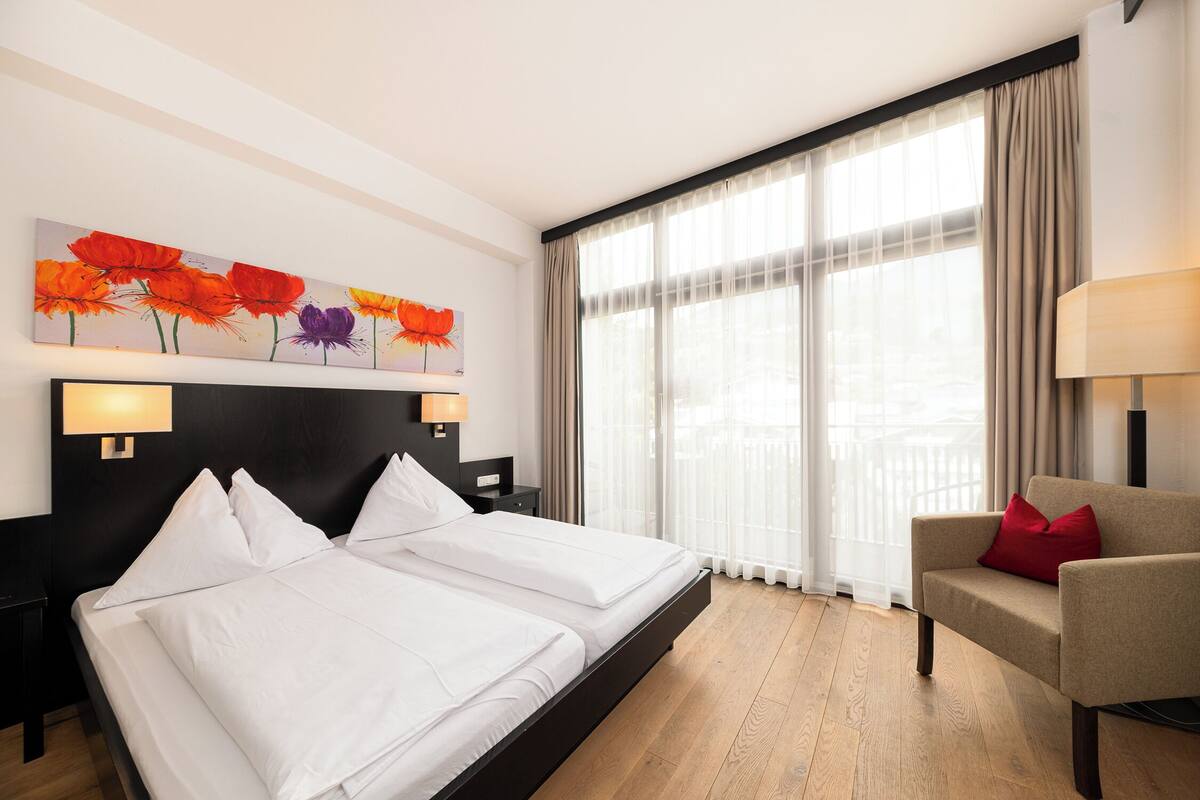 A hotel room with a double bed, a sofa, a painting on the wall, and a large window with curtains.