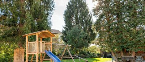 A wooden play structure with a slide and swings, surrounded by trees and a grassy yard.