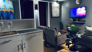 Ein modernes Wohnzimmer mit einer Couch, einem Esstisch und einem Fernseher. Auf einem Tablett stehen zwei Weingläser und es gibt einen Blumentopf.