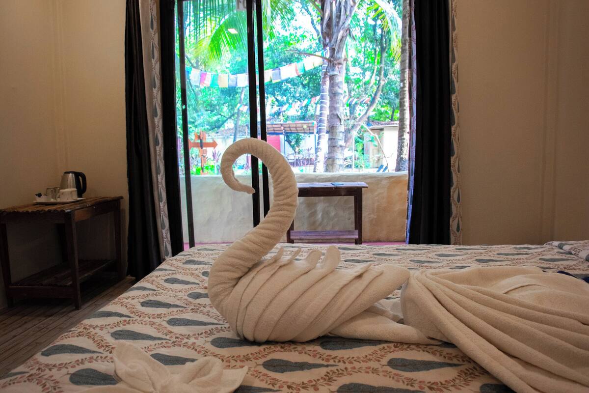 A bed with a swan-shaped towel, a coffee maker, and a view of a balcony with a palm tree.