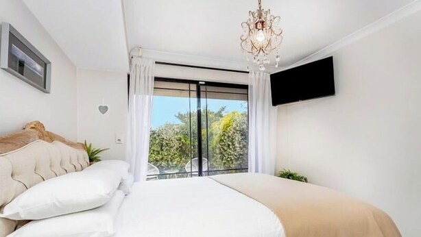 A bedroom with a large bed, a chandelier, a TV mounted on the wall, and a sliding glass door to the outside.
