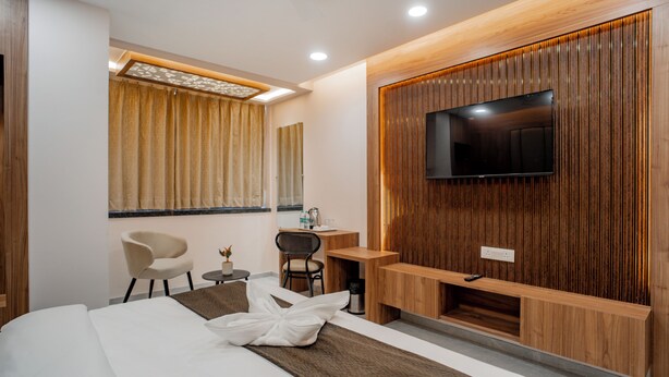 A modern hotel room with a large bed, a desk, a chair, a TV mounted on a wooden panel wall, and a window with curtains. - Hotel SK Elite (Jodhpur)