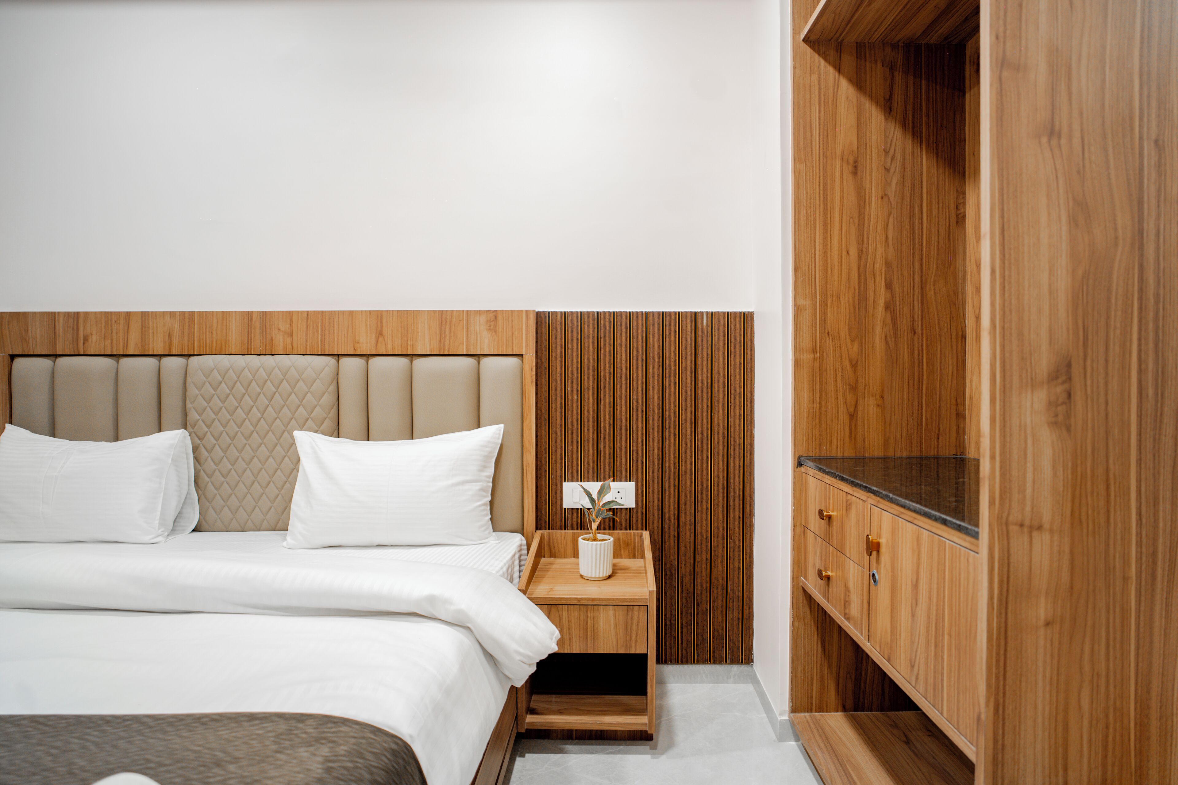 A modern hotel room with a wooden headboard, a wooden nightstand with a plant, and a wooden wardrobe.