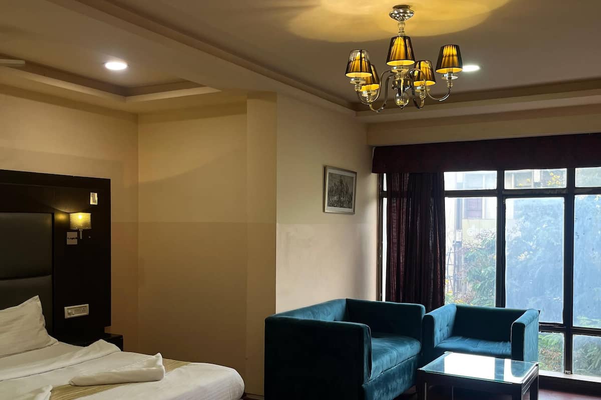Quarto de hotel com cama, sofá, mesa de vidro, lustre e uma grande janela com vista.