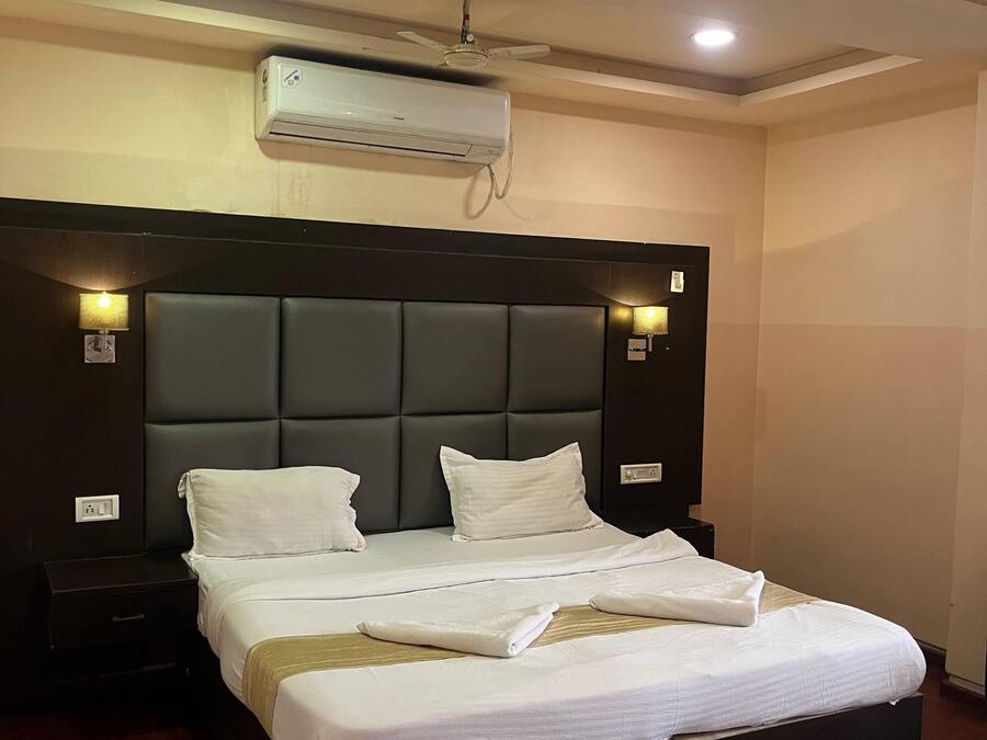 A hotel room with a large bed, dark headboard, bedside lamps, an air conditioning unit, and a ceiling fan.