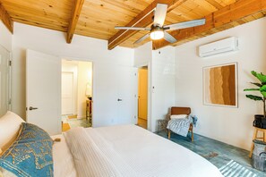 A bedroom with a bed, a ceiling fan, an air conditioning unit, a door, and a plant.