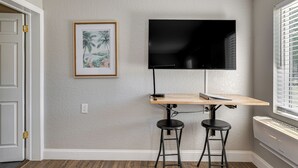 Une pièce avec un bureau en bois, deux tabourets de bar noirs, un téléviseur à écran plat fixé au mur et un tableau encadré accroché au mur.