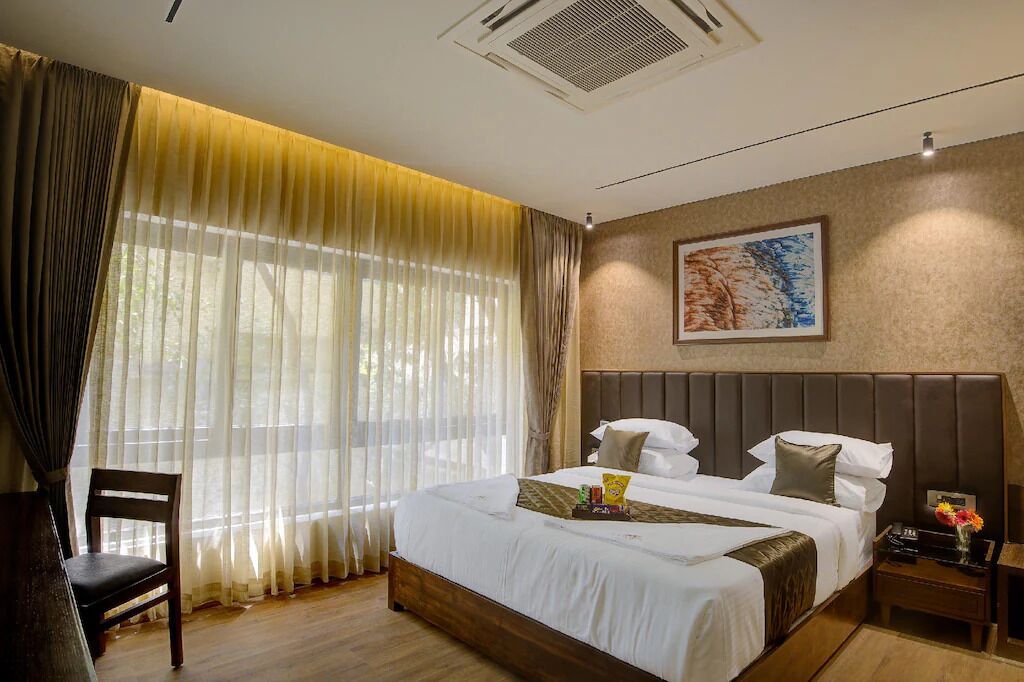 Quarto de hotel com uma cama grande, uma cadeira, uma mesa de cabeceira e uma janela com cortinas finas.