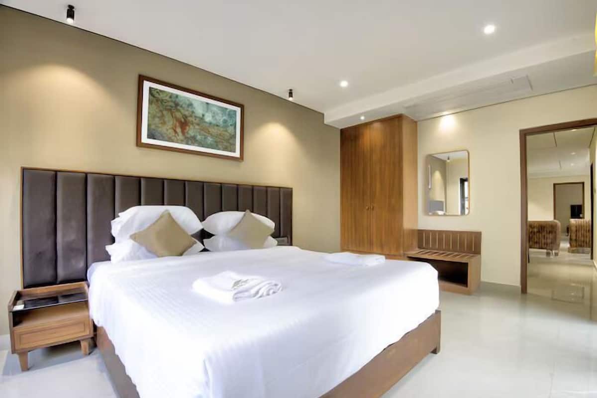 A modern hotel room with a large bed, wooden headboard, a nightstand, a wooden bench, a large mirror, and a painting on the wall.