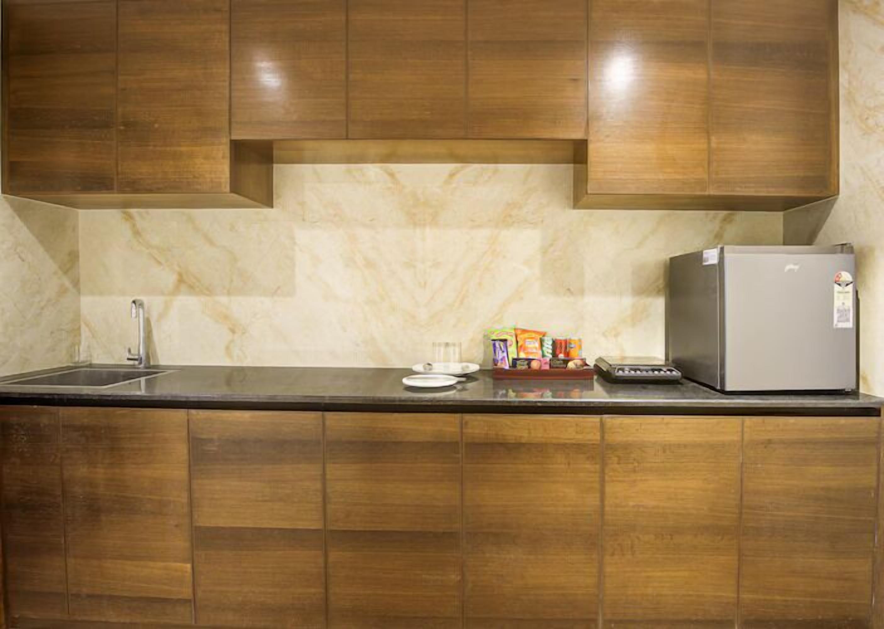 Una cocina con armarios de madera, salpicadero de mármol, pileta de acero inoxidable, refrigerador y un escurreplatos pequeño con alimentos.